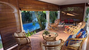 A covered outdoor seating area with wicker furniture, a small table with a plant, and a view of a swimming pool.