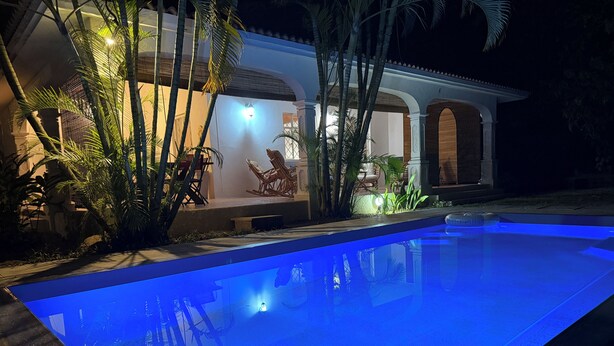 A swimming pool with a blue tint, surrounded by palm trees and a building with a patio area.