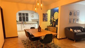A dining area with a wooden table and chairs, a kitchen in the background, and a sofa with cushions.