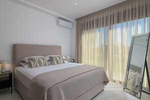 Een moderne slaapkamer met een groot bed, een nachtkastje met lamp, een spiegel en dunne gordijnen.