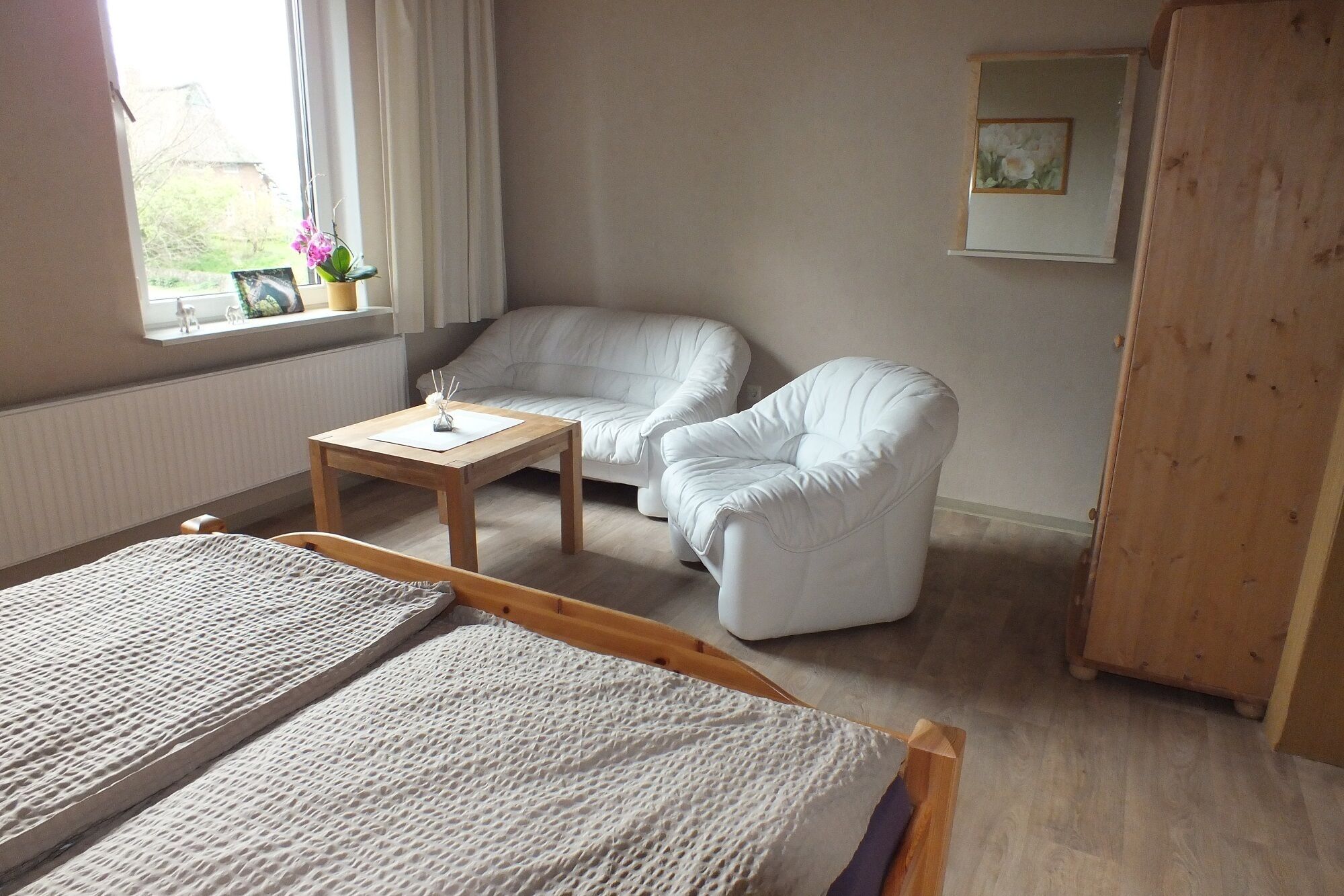 Una habitación con una cama, dos sillones blancos, una mesita de madera, una ventana con cortinas y un armario de madera.