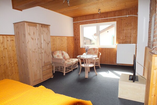 Una habitación con cama, armario de madera, sillas de mimbre, una mesita redonda y un televisor sobre un mueble.