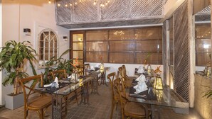Comedor con sillas y mesas de madera, ventanas amplias y plantas decorativas.
