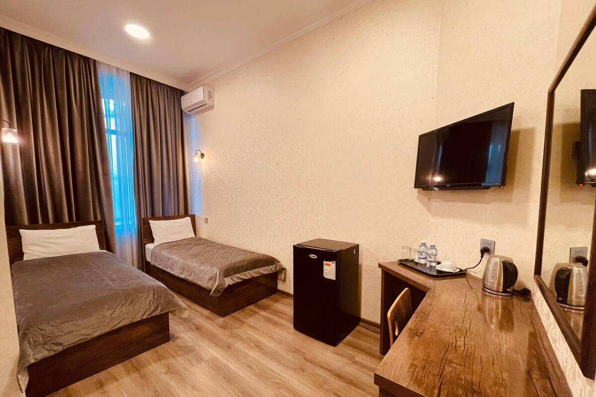A hotel room with two beds, a desk with a mirror, a TV, and a small kitchenette.