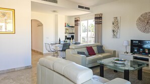 Un salon moderne avec un canapé, une table basse en verre et un téléviseur. On aperçoit à l’arrière-plan un coin repas avec une table et des chaises.