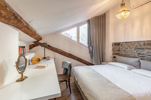 A bedroom with a sloped ceiling, exposed wooden beams, a stone wall, a bed with white bedding, a desk with a lamp, and a hanging chandelier. - Como Dreamhouse (Como)