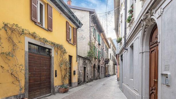 A narrow cobblestone street flanked by historic buildings with traditional shutters and a mix of stone and plaster facades.