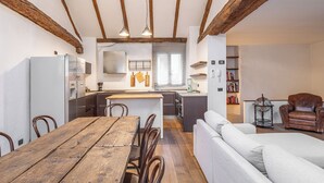 A cozy kitchen with wooden beams, a dining table with chairs, a sofa, and a small bookshelf.