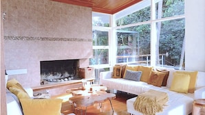 A cozy living room with a fireplace, large windows, and a white sofa with yellow cushions.