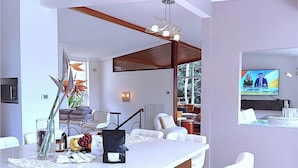 A modern living room with a white sofa set, a dining area with a white table, and a television mounted on the wall.