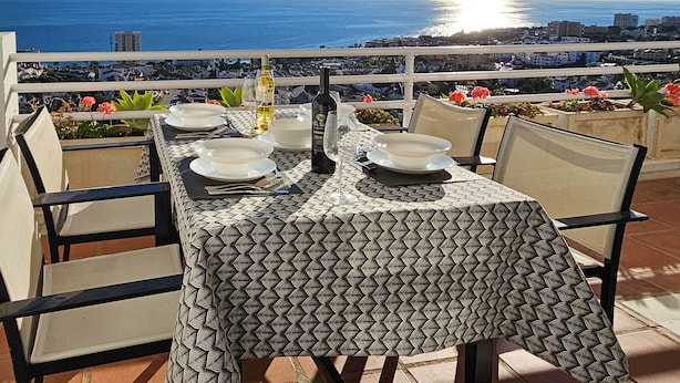 A table set for two with a patterned tablecloth, overlooking a coastal cityscape with a clear sky and the sun setting over the sea. - Two bedroom apartment in Riviera del Sol (Mijas Costa)