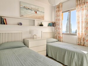 Slaapkamer met twee bedden, een nachtkastje, een lamp, een plank met boeken, een schilderij en een raam met gordijnen.