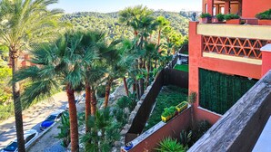 Un hôtel à la façade rouge, doté d’un balcon offrant une vue sur des palmiers et un paysage vallonné, ainsi que d’un parking où sont garées des voitures.