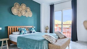 Une chambre à coucher comprenant un lit, un banc en osier, un tabouret en bois et donnant sur un espace extérieur aménagé avec des tables et des chaises.