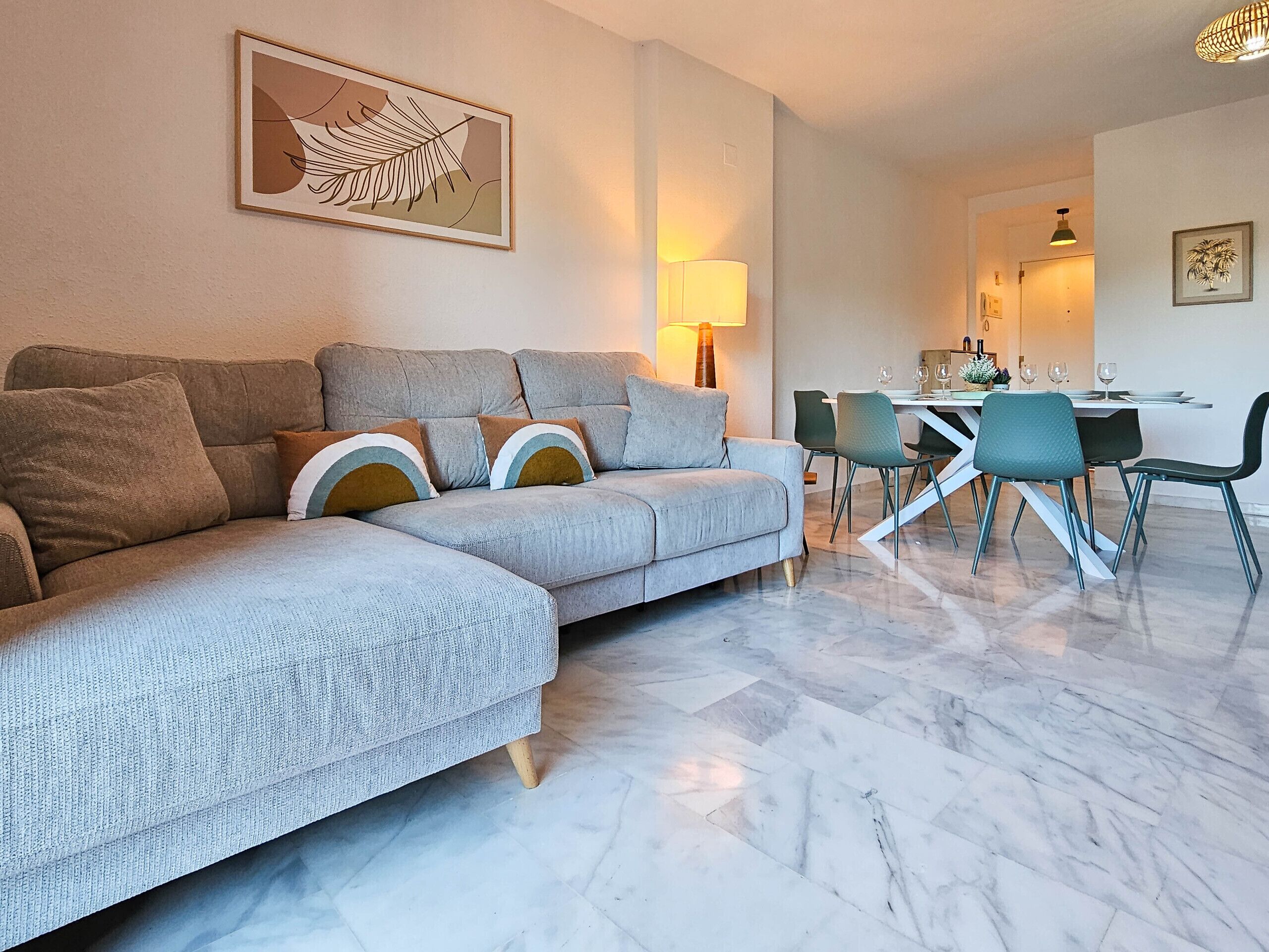 A living room with a grey sectional sofa, a dining table with chairs, a lamp, and a framed artwork on the wall.