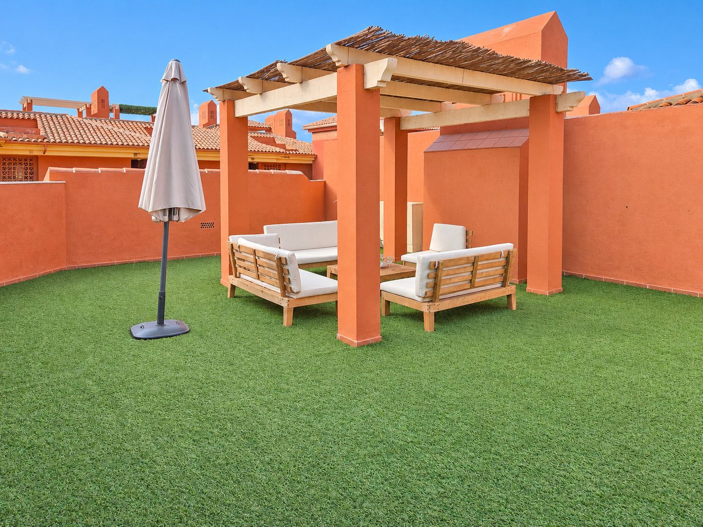 A patio with a wooden pergola, two white cushioned chairs, and a single umbrella.