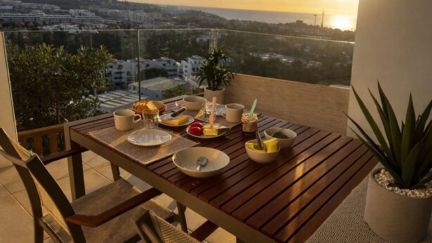 A balcony with a wooden table set for breakfast, overlooking a cityscape at sunset. - Three bedroom apartment in La Cala de Mijas (La Cala de Mijas)