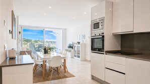 A modern kitchen with white cabinetry, a dining area with a table and chairs, and a large window offering a city view. - Three bedroom apartment in La Cala de Mijas (La Cala de Mijas)