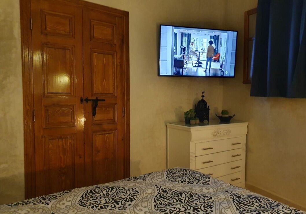 A bedroom with a bed, a wooden wardrobe, a television mounted on the wall, and a dresser with drawers.