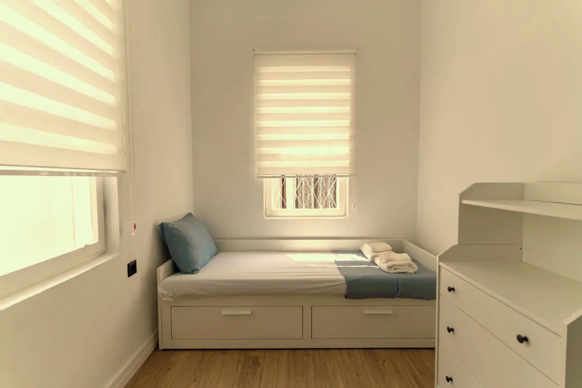 A small, bright room with a single bed, a window with blinds, and a white dresser.