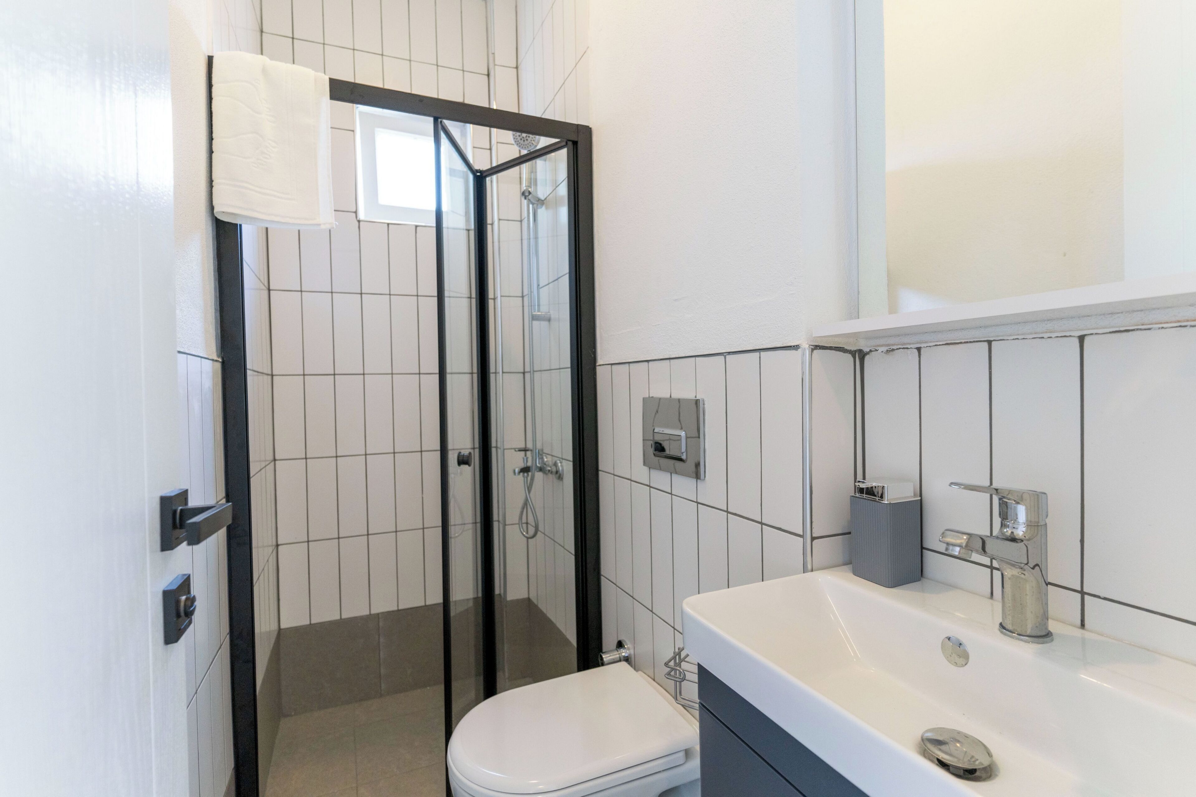 Egy modern fürőszoba, üvegfalas zuhanyzóval, fehér WC-vel és egy mosdókagylóval, mely felett tükör található.