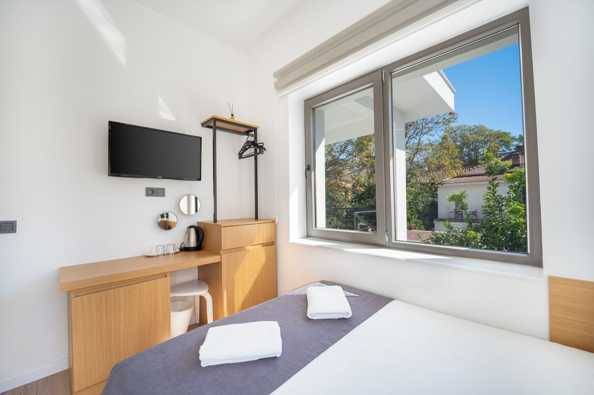 Une chambre d’hôtel moderne équipée d’un lit, d’un bureau, d’une télévision et d’une fenêtre donnant sur des arbres et des immeubles.
