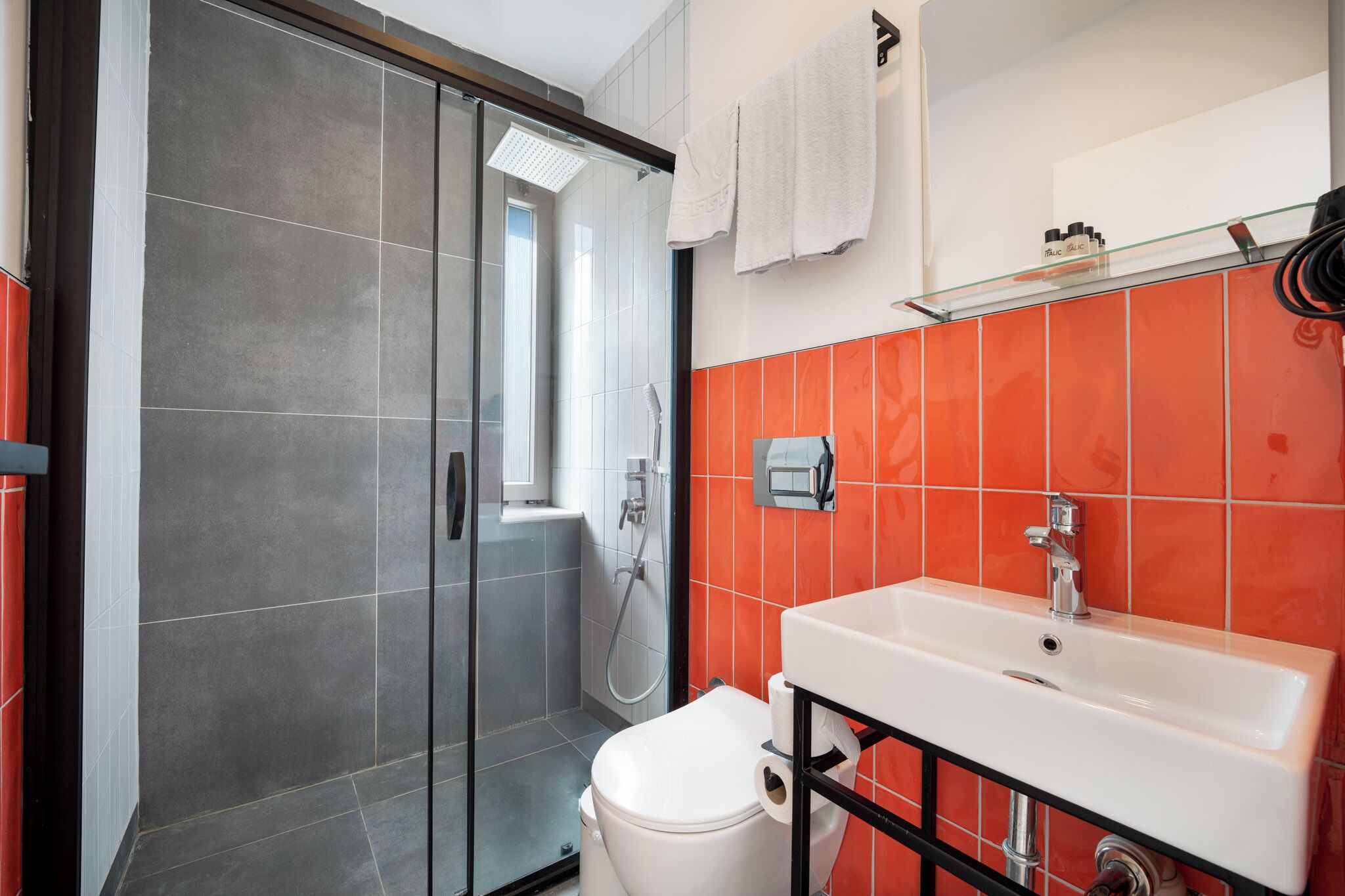 A modern bathroom with a glass shower enclosure, a white toilet, and a sink with a red-tiled backsplash.