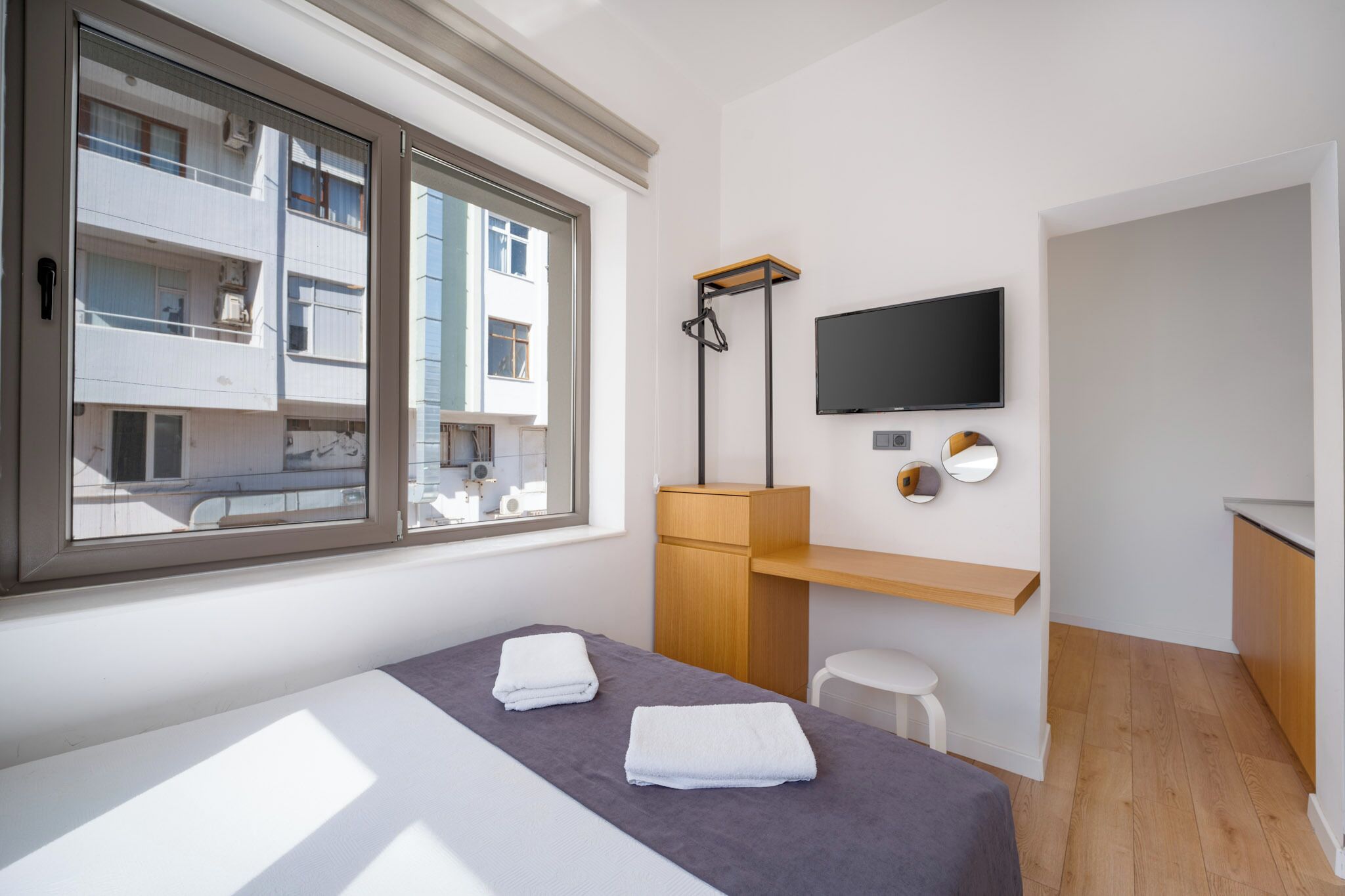 A modern hotel room with a large window, a bed with towels, a wooden desk, a flat-screen TV, and a small stool.