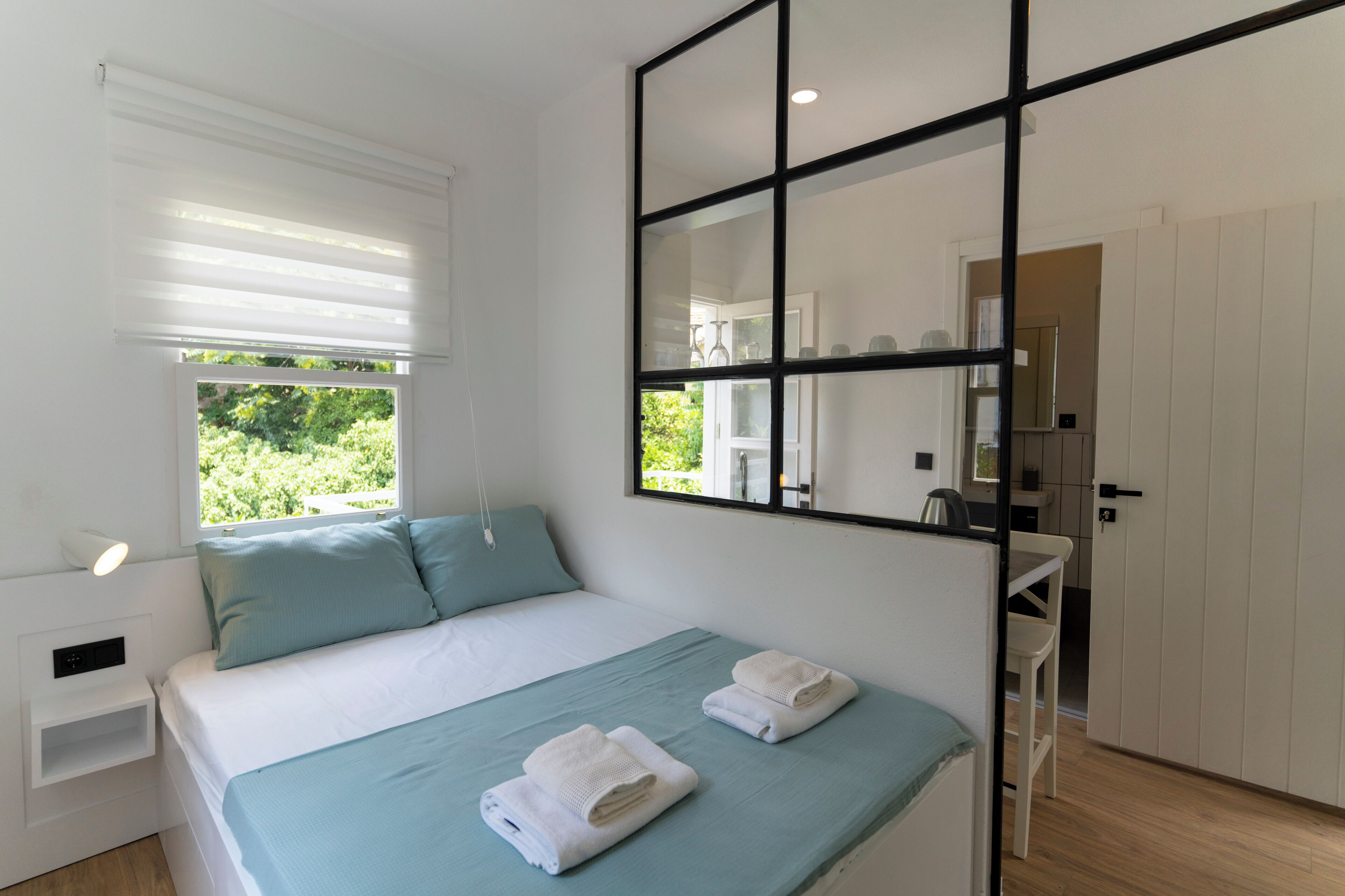 A modern bedroom with a bed, a bedside table, a window with blinds, a mirror, and a view of the outdoors.