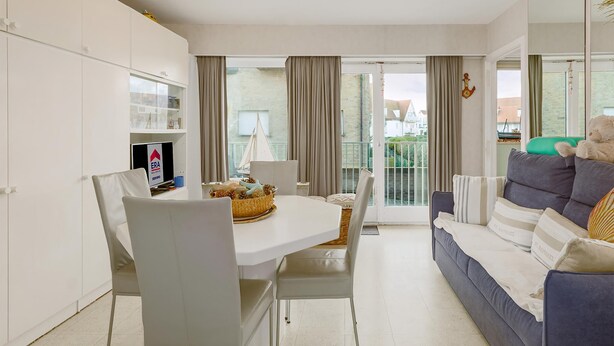 Een moderne woonkamer met een eethoek, witte meubels, een bank met kussens en uitzicht op buiten.