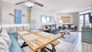 Een moderne woonkamer met een houten eettafel, banken, een plafondventilator en uitzicht op de stad door grote ramen.