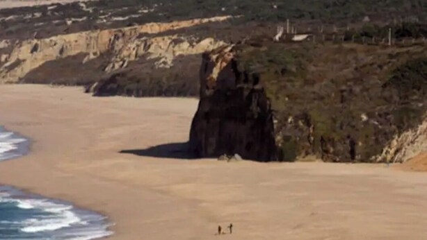 A beach with a rocky cliff and a few people walking along the shore.