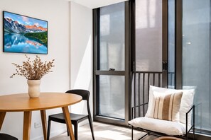 A modern room with a wooden table, a black chair, a sofa with a striped pillow, and a large window with a view of mountains. - Stylish 1BR | Pool + Gym | Courtenay (Wellington)
