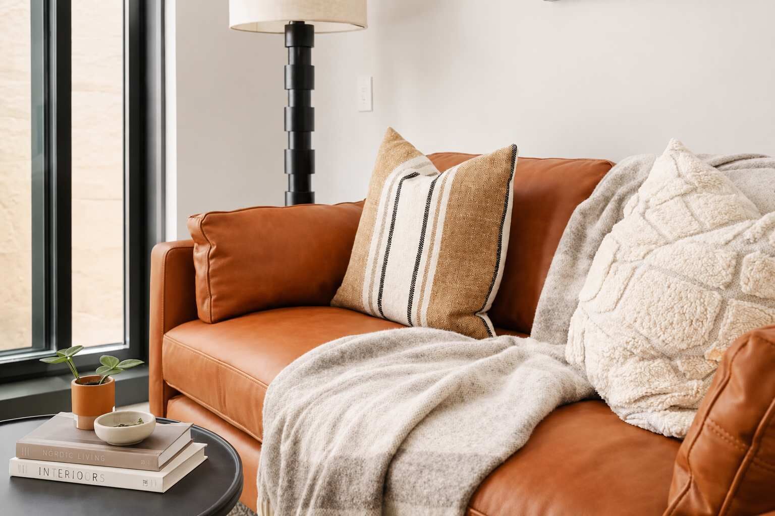 A brown leather sofa with a white throw blanket and striped pillows, a small table with books and a plant, and a floor lamp.