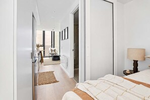 A modern hotel room with a bed, bedside table, lamp, and a view of the hallway leading to a bathroom.