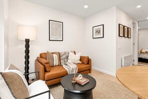 A modern living room with a brown leather sofa, a round black coffee table, and a floor lamp. There are framed artworks on the walls and a door leading to another room. - Stylish 1BR | Pool + Gym | Courtenay (Wellington)