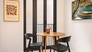 Una habitación con una mesa redonda de madera, dos sillas negras, un cuadro enmarcado en la pared y una ventana con persianas verticales.