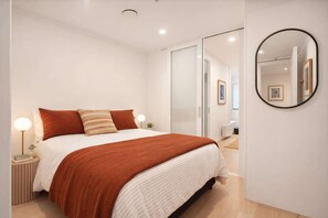 A modern bedroom with a bed, bedside lamps, a mirror, and a door leading to another room.