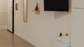 Un corridoio con una porta, uno specchio, un quadro incorniciato, una mensola con oggetti decorativi, una televisione e un frigorifero.