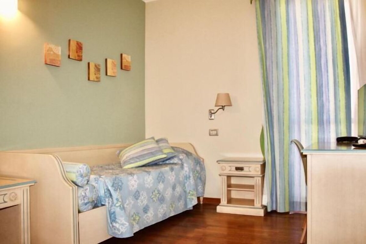 Een kamer met een bed, nachtkastje, lamp en raam met gestreept gordijn.