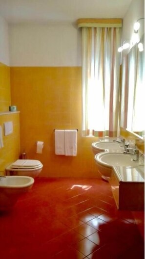 Un baño con paredes amarillas, inodoro blanco, bidé, lavamanos y espejo con luces.