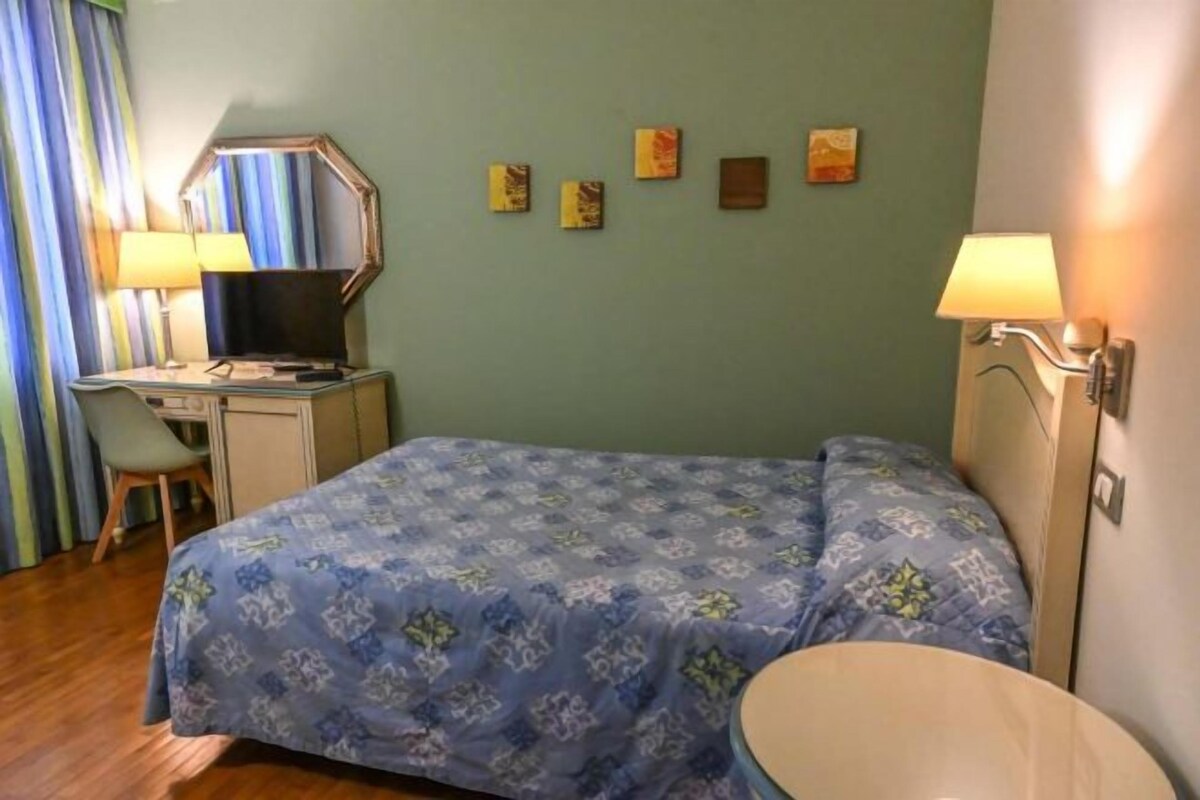 Quarto de hotel com cama, escrivaninha, cadeira e uma pequena mesa redonda.