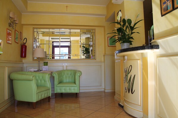 A room with two green armchairs, a mirror, a potted plant, and a cream-colored cabinet with a monogram.