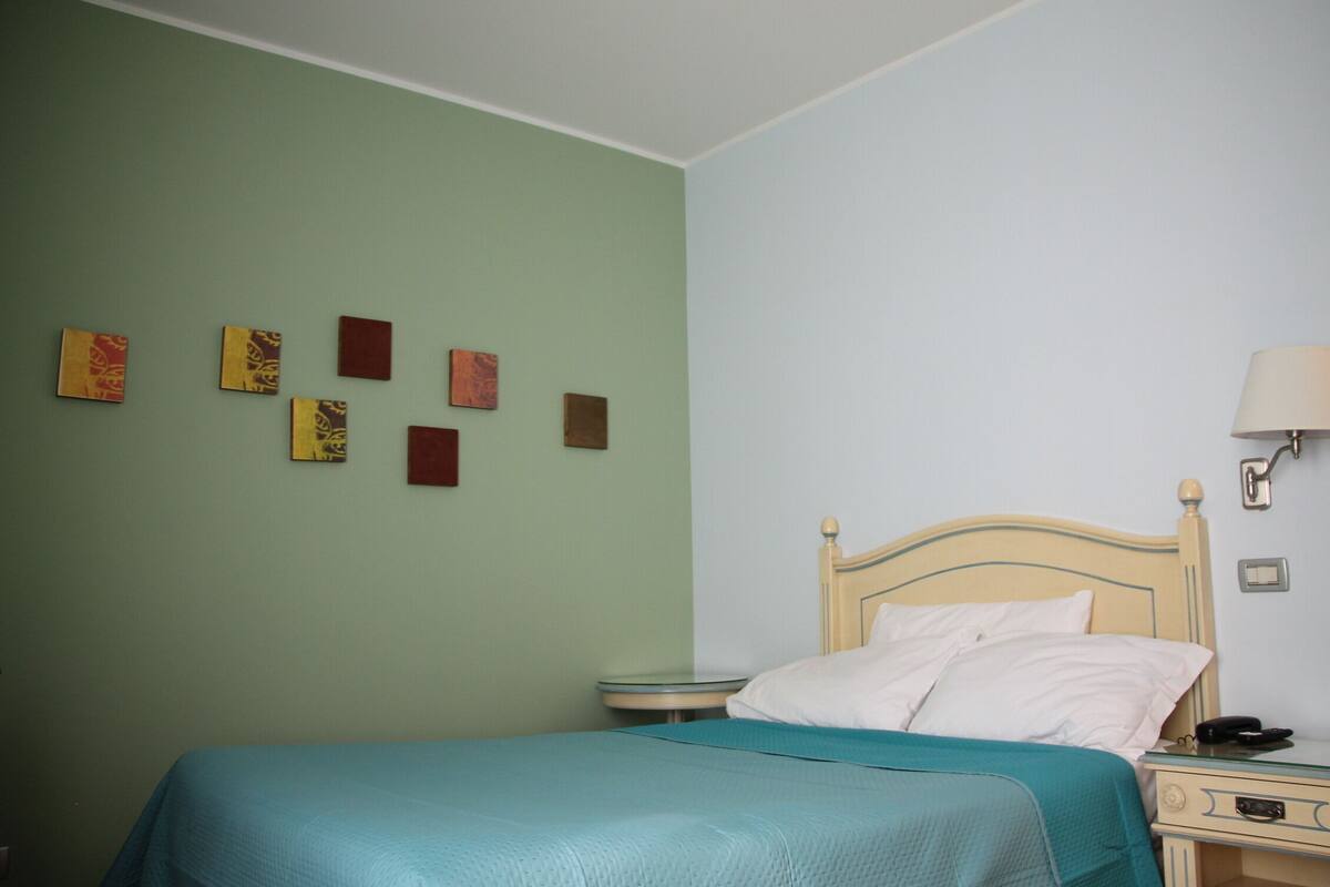 A single bed with a teal bedspread, a wooden headboard, a bedside table with a lamp, and a wall with several framed artworks.