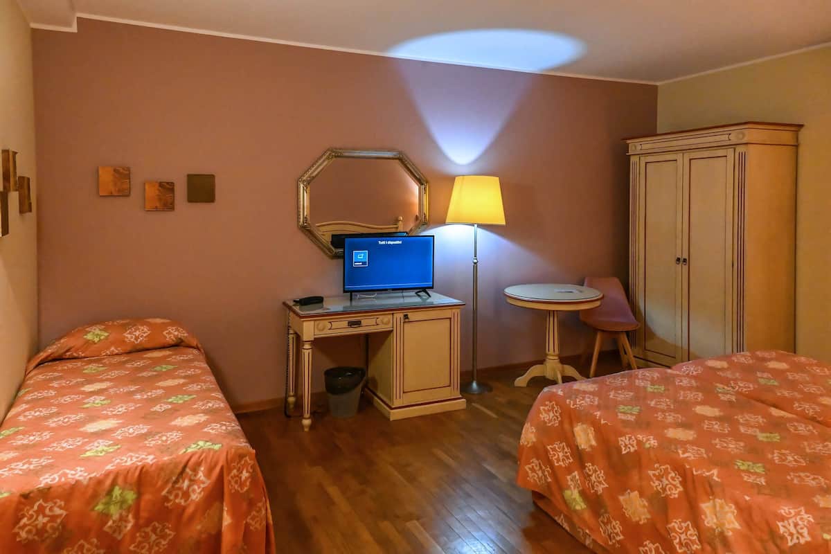 A hotel room with two beds, a desk with a computer, a lamp, a small table, and a wardrobe.