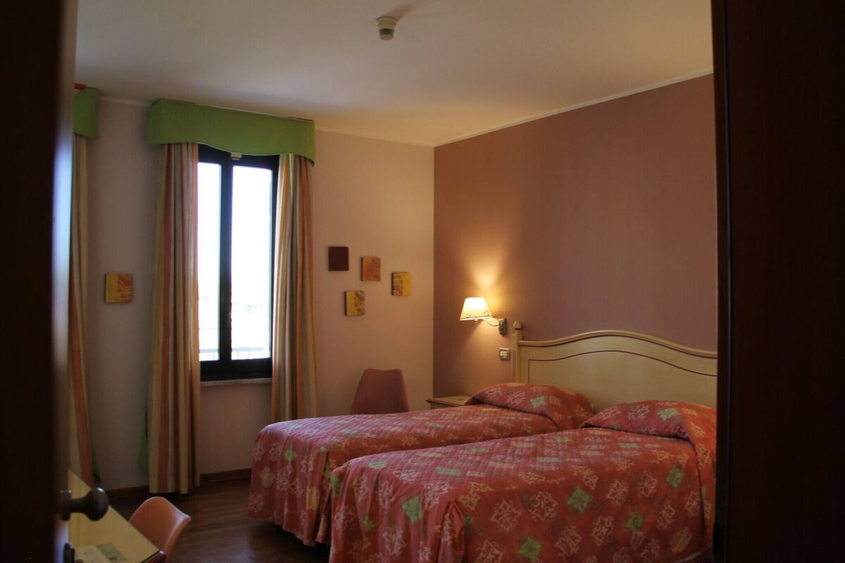 A hotel room with two beds, a chair, a window with curtains, a wall-mounted lamp, and a door.