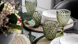 A dining table set with green glassware, white plates, and woven placemats.