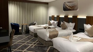A hotel room with three beds, a desk, a chair, and a large window with curtains. - How inn Hotel (Madinah)