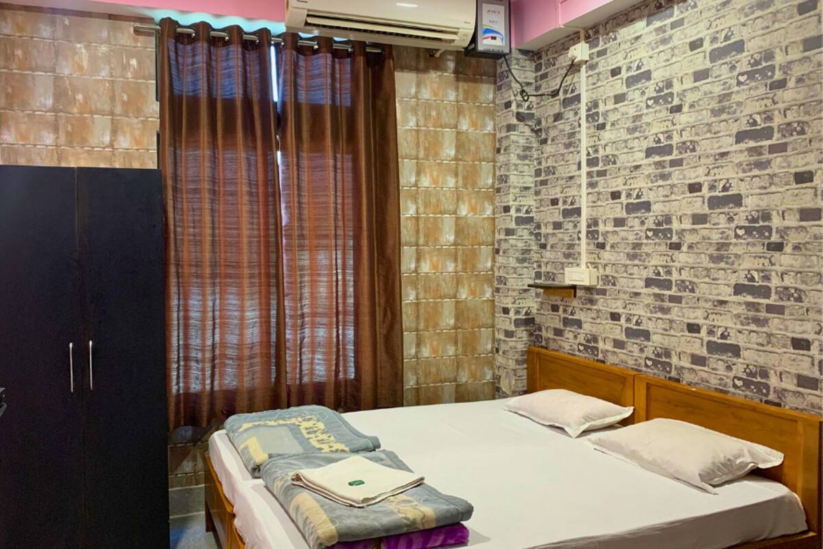A bedroom with a brick-patterned wall, a wooden bed, a dark wardrobe, and a window with brown curtains.
