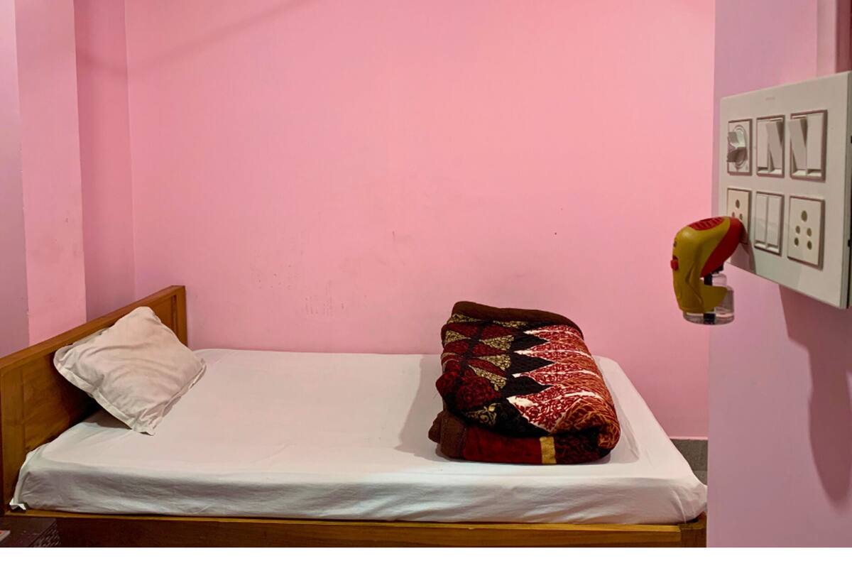 A single bed with a white mattress and a wooden headboard, a white pillow, and a red and black patterned blanket.